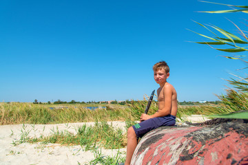 A boy is playing on a black clarinet sitting on an old wooden boat on the beach.