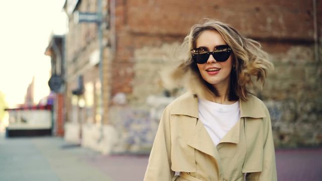 Slow motion portrait of attractive blond girl walking in the street, smiling and looking at camera. Cheerful people, beautiful clothes and urban lifestyle concept.
