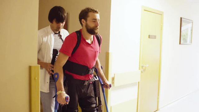 Young disable man walking in the robotic exoskeleton in the rehabilitation clinic. Doctor supports him
