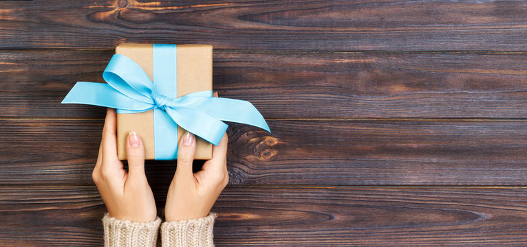 Woman hands give wrapped valentine or other holiday handmade present in paper with blue ribbon. Present box, decoration of gift on white wooden table, top view with copy space. Banner
