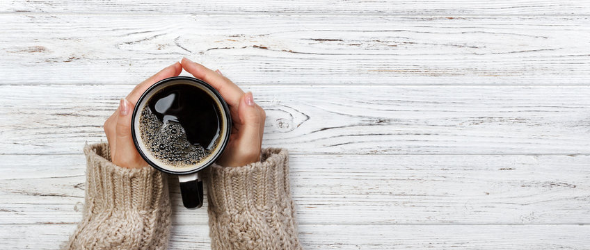 Woman holding cup of hot coffee on rustic wooden table, closeup photo of hands in warm sweater with mug, winter morning concept, top view. Banner