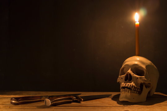 Human skull with candle light and knife on wooden table in the dark background, Decorate for Halloween Theme with copy space.