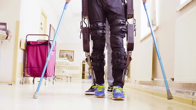 Close up legs of disable man in the robotic exoskeleton walking through the corridor with support of a doctor. Then camera moving to the faces. Both smiling.