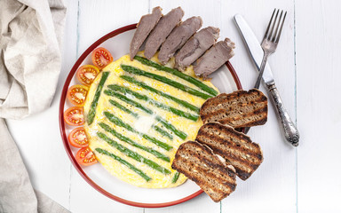 Omelette with asparagus. Homemade ham, cherry tomatoes, toast. Wooden light background. Top view
