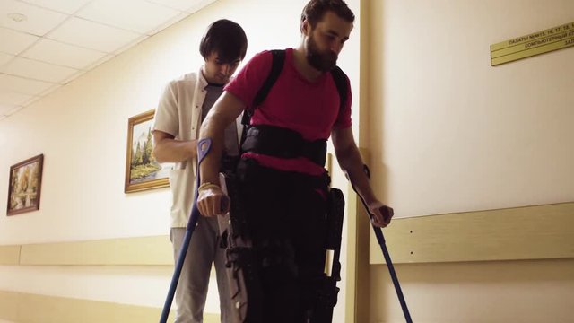 Young disable man walking in the robotic exoskeleton in the rehabilitation clinic with support of the doctor.
