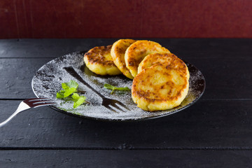 Cottage cheese pancakes on a dark background. Traditional Ukrainian dish - syrniki with fresh mint. Pancakes with cottage cheese on a black plate sprinkled with powdered sugar