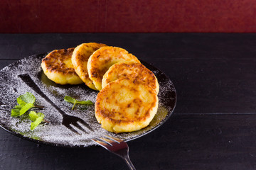 Cottage cheese pancakes on a dark background. Traditional Ukrainian dish - syrniki with fresh mint. Pancakes with cottage cheese on a black plate sprinkled with powdered sugar