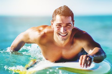 Handsome young surfer floating in blue water - obrazy, fototapety, plakaty