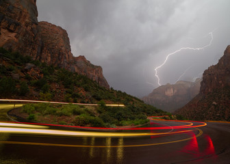 Lightning lights the sky and canyon as a car leaves trails of red and yellow on a switchback bend in the road during an early morning summer monsoon storm in Zion National Park.