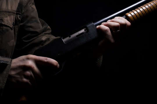 The man keeps a pump shotgun in his hand in Finland. He is wearing a camouflage jacket. Photo taken in a studio. Image includes strong shadows.