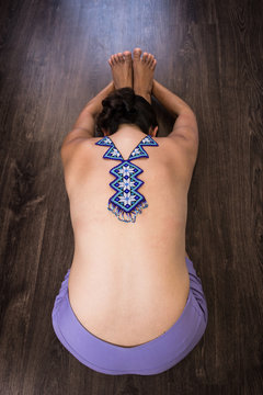 Top less woman on seated forward bend pose with huichol blue necklace on naked back. Female yogi on paschimottanasana on wood planks floor. Fashion design, Mexican handcraft, decoration art concepts
