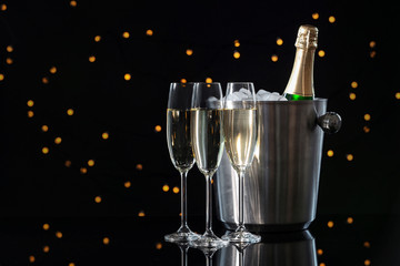 Glasses with champagne and bottle in bucket on dark table against blurred lights