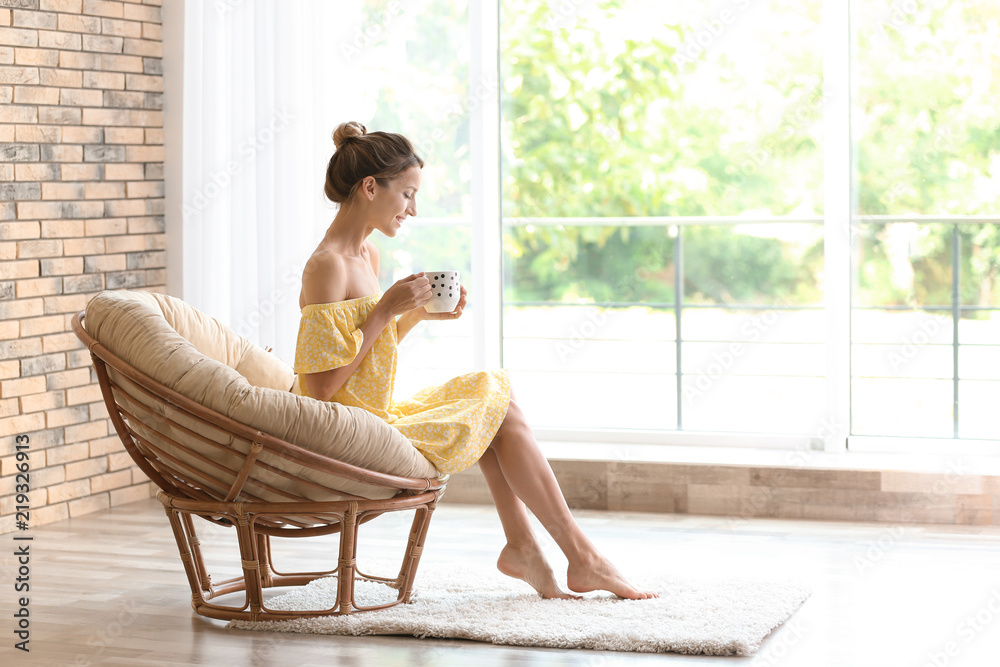 Canvas Prints Young woman with cup of aromatic coffee sitting in papasan chair near window at home