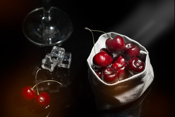 View of sweet cherry in sack among glass and ice cube in black background with flare and foreground.