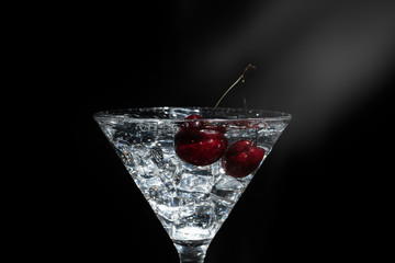 Close up view of splash water with falling cherry in a martini glass among ice in black background with flare.