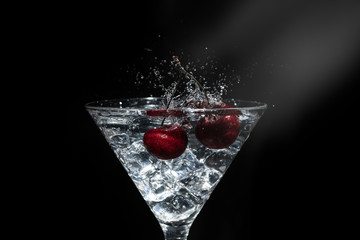 Close up view of splash water with falling cherry in a martini glass among ice in black background with flare.