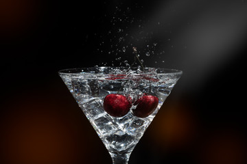 Close up view of splash water with falling cherry in a martini glass among ice in black background with flare and foreground.