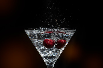 Close up view of splash water with falling cherry in a martini glass among ice in black background foreground.
