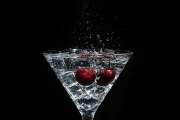 Close up view of splash water with falling cherry in a martini glass among ice in black background.