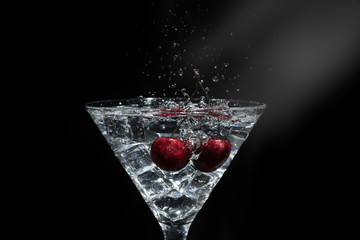 Close up view of splash water with falling cherry in a martini glass among ice in black background with flare.