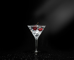 Close up view of splash water with falling cherry in a martini glass among ice in black background with flare.