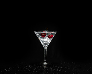 Close up view of splash water with falling cherry in a martini glass among ice in black background.