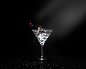 Close up view of splash water with falling cherry in a martini glass among ice in black background with flare.
