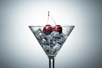 Close up view of splash water with falling cherry in a martini glass among ice in white background.