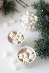 Cocoa with marshmelow in white cups and marshmallows in a glass jar on a white table with spruce branches and garland.