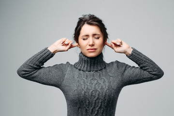 Waist up portrait of angry young girl blocking ears with fingers and closing eyes. Unhappy and annoyed female in stylish sweater. Isolated on gray background