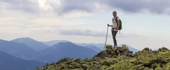 Foto op Aluminium Young slim blond tourist girl with backpack and stick standing on rocky top against bright blue morning sky on foggy mountain range panorama background. Tourism, traveling and climbing concept. © bilanol