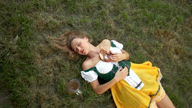 Beautiful drunk young lady in a traditional Bavarian dress lies on the ground. Music festival oktoberfest. Near it stands an empty glass of beer and pretzel. On the lips of a woman red lipstick