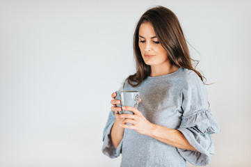 Beautiful woman holding cup of coffee or tea, wearing grey dress, posing against white background