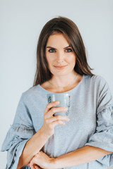 Beautiful woman holding cup of coffee or tea, wearing grey dress, posing against white background