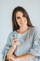 Beautiful woman holding cup of coffee or tea, wearing grey dress, posing against white background