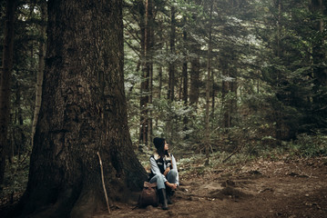 hipster traveler with backpack sitting under tree in woods. stylish woman hiking. wanderlust and travel concept with space for text. atmospheric moment