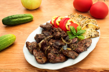 Plate with stewed liver and apples, slices of pepper, tomatoes and a sprig of basil, next to a wooden background cucumbers, tomatoes and apple.
