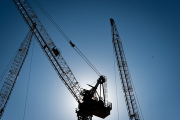 Three construction cranes in silhouette with the sun directly behind and a blue sky, horizontal