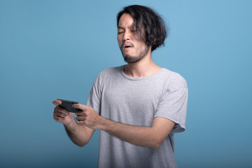 Upset Young bearded man playing mobile games in blue background.