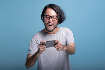 Excited Young bearded man playing mobile games in blue background.