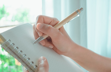 Woman hand is writing on white notebook with nature light from window and white curtain in background.
