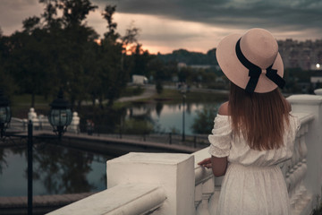 Fototapeta premium A beautiful young girl in a long white dress and hat walks through the evening park with a pond in the rays of the sunset