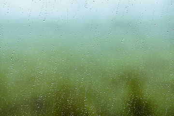 Dirty glass with drops of rain. Raindrops on green and blue clear background. Greenery and sky in bokeh outside window. Droplets and stains close up. Detailed transparent texture in macro.
