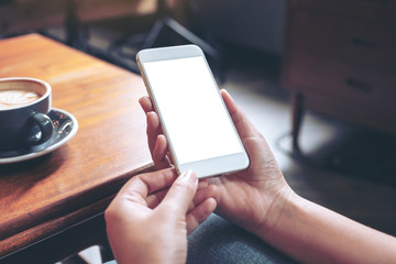 Mockup image of hand holding white mobile phone with blank desktop screen with coffee cup on wooden table in cafe