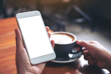 Mockup image of hands holding white mobile phone with blank screen while drinking coffee in modern cafe