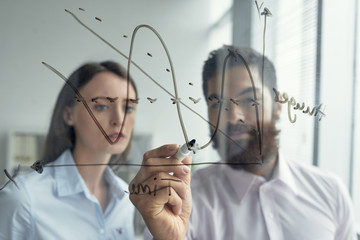 Businessman drawing chart on glass wall to explain his idea to coworker