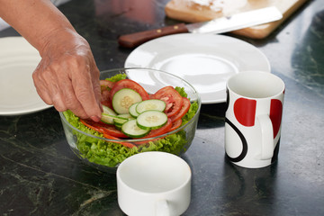 Hand of woman putting cut cucumber into bowl of salad