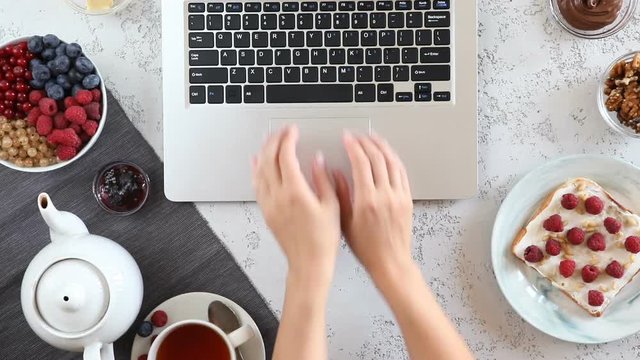 Top view of woman hands on workspace with coffee break with laptop, bread toasts with berries and cup of black tea composition. Flat lay, view from above