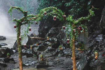 Tropical wedding ceremony with waterfall view in jungle canyon. Decorated with green ivy, old branches and hanging lamps