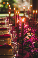 Wooden wedding table decorated with red candles, pink cloth and purple orchids. Romantic family dinner in evening